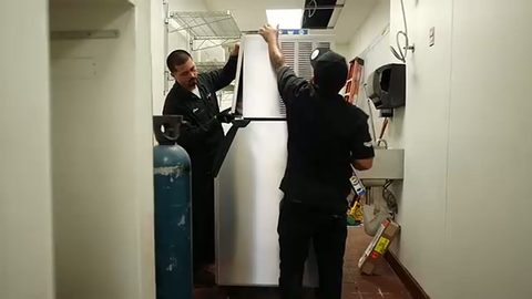 two movers standing up a stainless commercial refrigerator in a restaurant, face visible