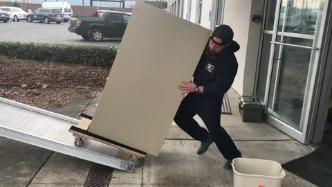 Professional mover wheeling a large desktop panel on a four-wheel furniture dolly into a building, face visible