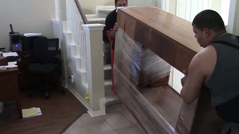 Two movers carrying a shrink-wrapped wooden entertainment center up an interior staircase with a red lifting strap, face visible