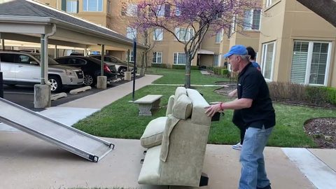 Two movers carrying a sofa across a residential driveway toward the moving truck, face visible