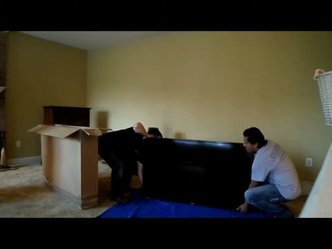 Two-man moving crew lowering a large flat-screen TV onto a blue moving blanket on the floor, one kneeling, face visible